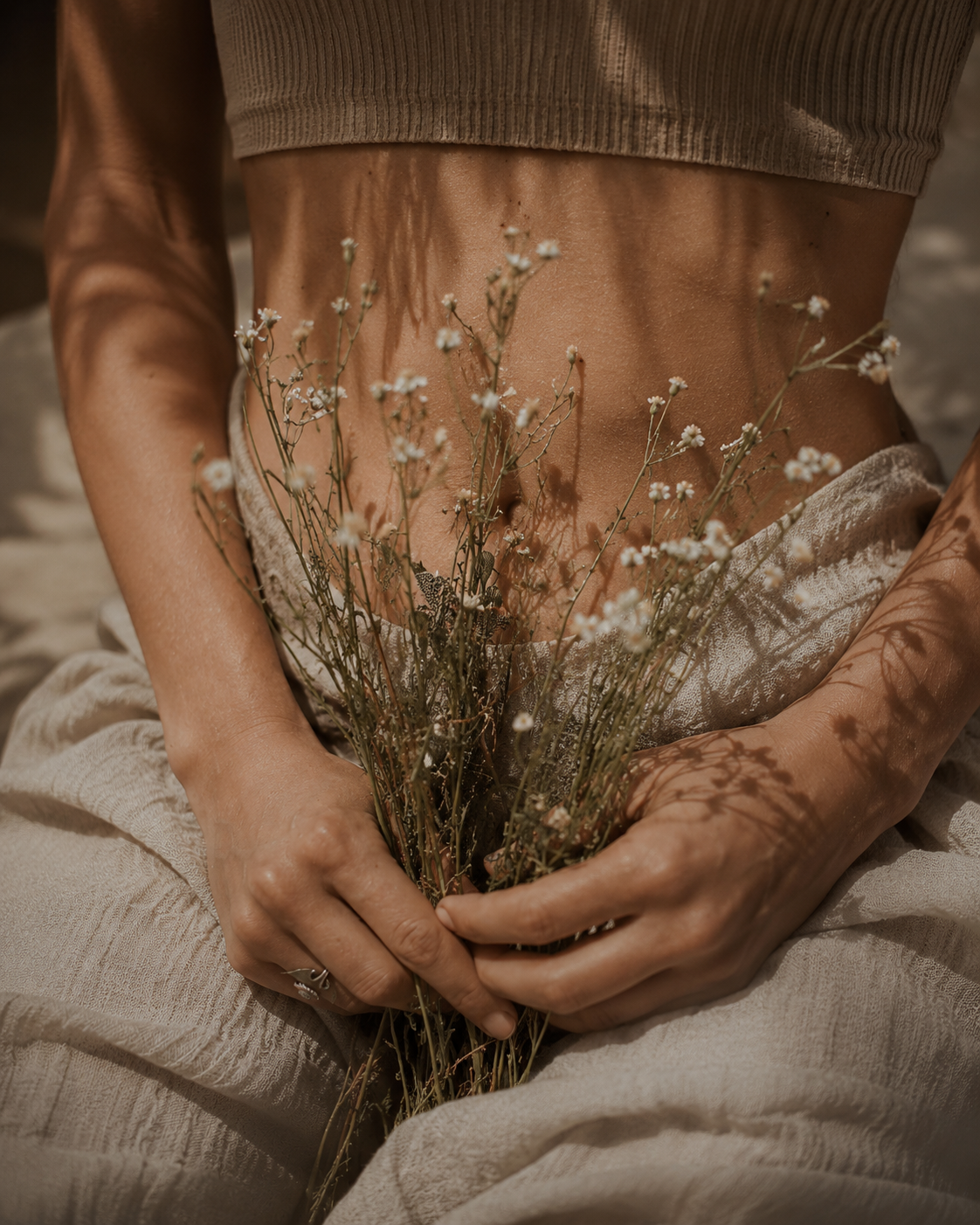 A woman holding wildflowers at her womb space, draped in linen
